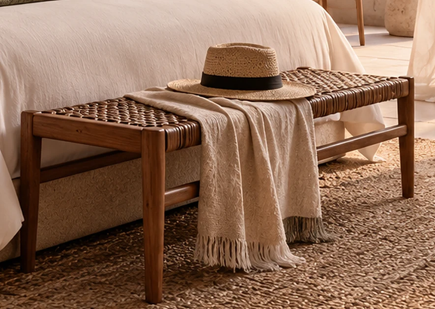 A woven bench with a straw hat and a folded throw on a jute rug at the foot of a bed